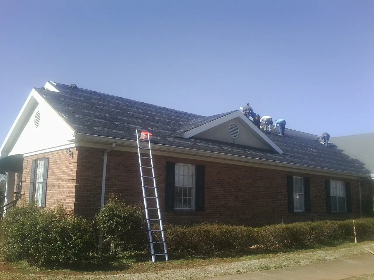 Crew tearing shingles off a brick ranch roof with a ladder set — Emergency Roof Repair work by our crew in Rocky Point